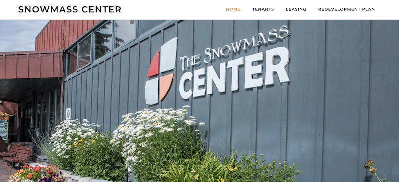 Hero image for The Snowmass Center website, featuring the center's logo on the side of a gray building. The outer wall is lined with blooming daisies.